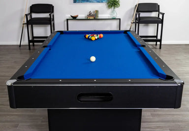 Full end view of the Hathaway Hustler 7-foot pool table with blue felt, racked billiard balls, and cue ball. Modern game room setting with two chairs, a shelf, and decor in the background.