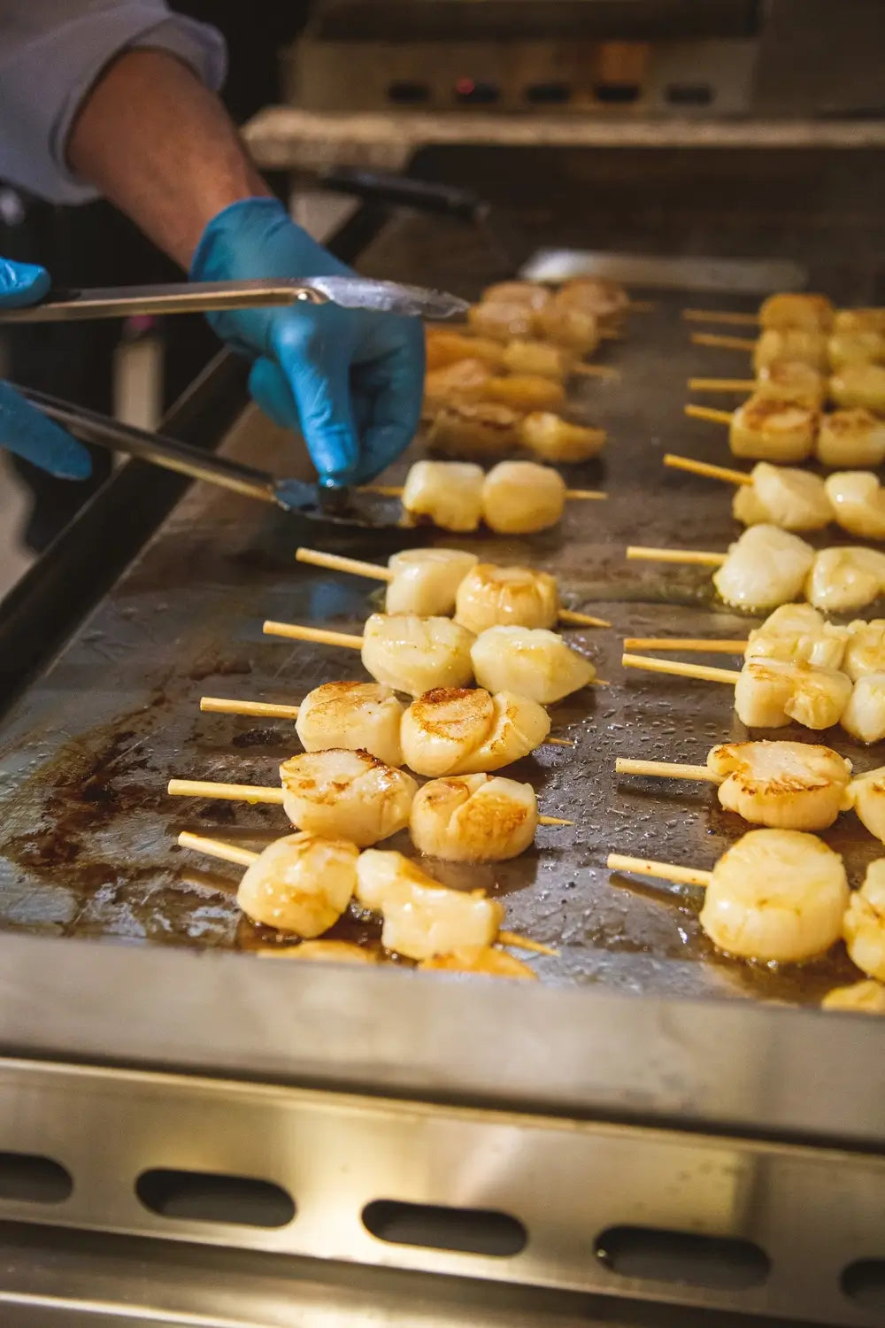 Chef grilling scallops on skewers over stainless steel Le Griddle cooktop.