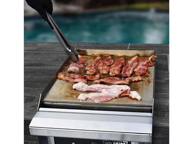 Wildfire Ranch 15″ Black Built-In Side Griddle – 17,000 BTUs actively cooking strips of bacon on a stainless steel griddle surface beside a poolside patio, demonstrating efficient heat distribution and grilling performance.
