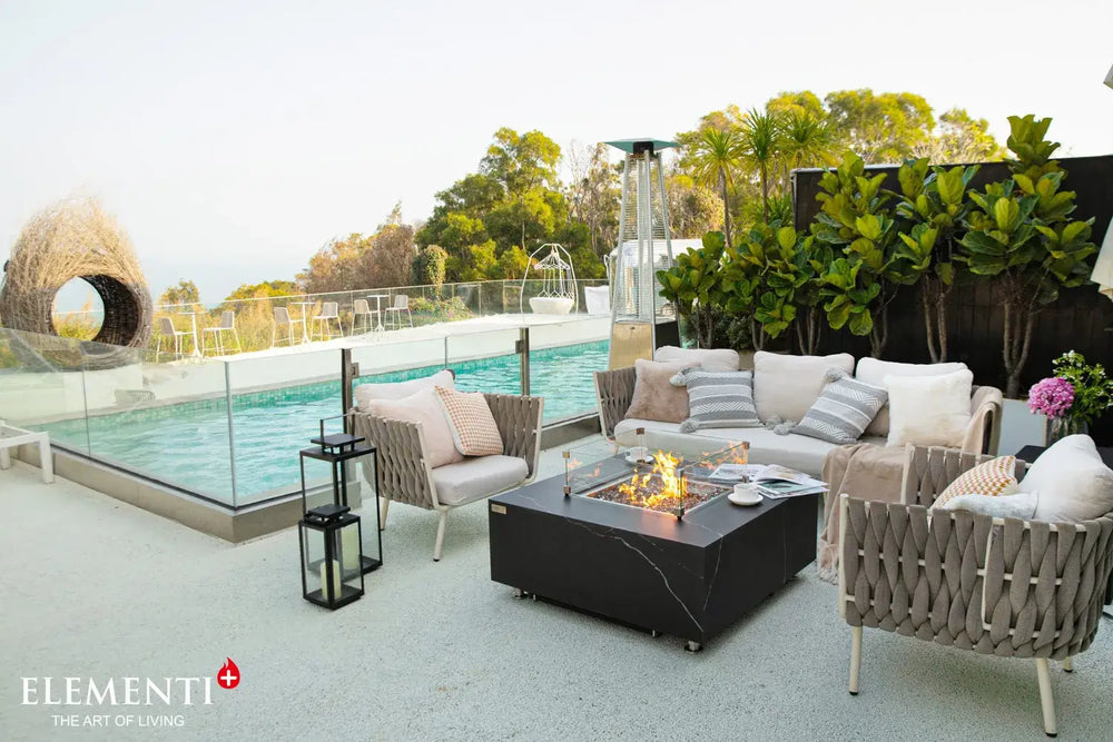 Modern outdoor living area featuring the Bulgaria Black rectangular gas fire pit table, a designer woven bird's nest chair, and a classic silver trailer in a poolside setting.