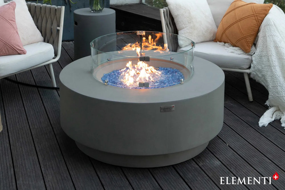 Elementi Plus Colosseo fire table with glass wind guard, blue fire glass, and flame on outdoor deck.