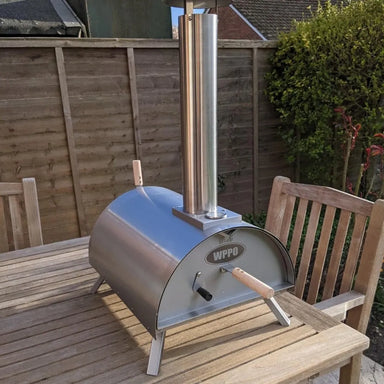 WPPO Lil Luigi portable wood-fired oven on outdoor wooden table with stainless chimney, folding legs, and rear handle.
