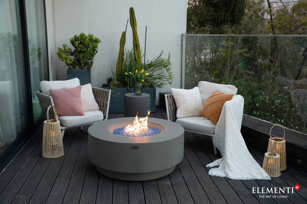 Elementi Plus Colosseo light gray fire table on balcony patio with lounge chairs and blue glass flame.