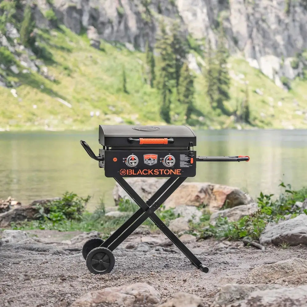 Foldable Blackstone 22" griddle with dual burners and hood set up near mountain lake for camping