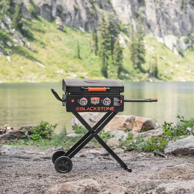 Foldable Blackstone 22" griddle with dual burners and hood set up near mountain lake for camping