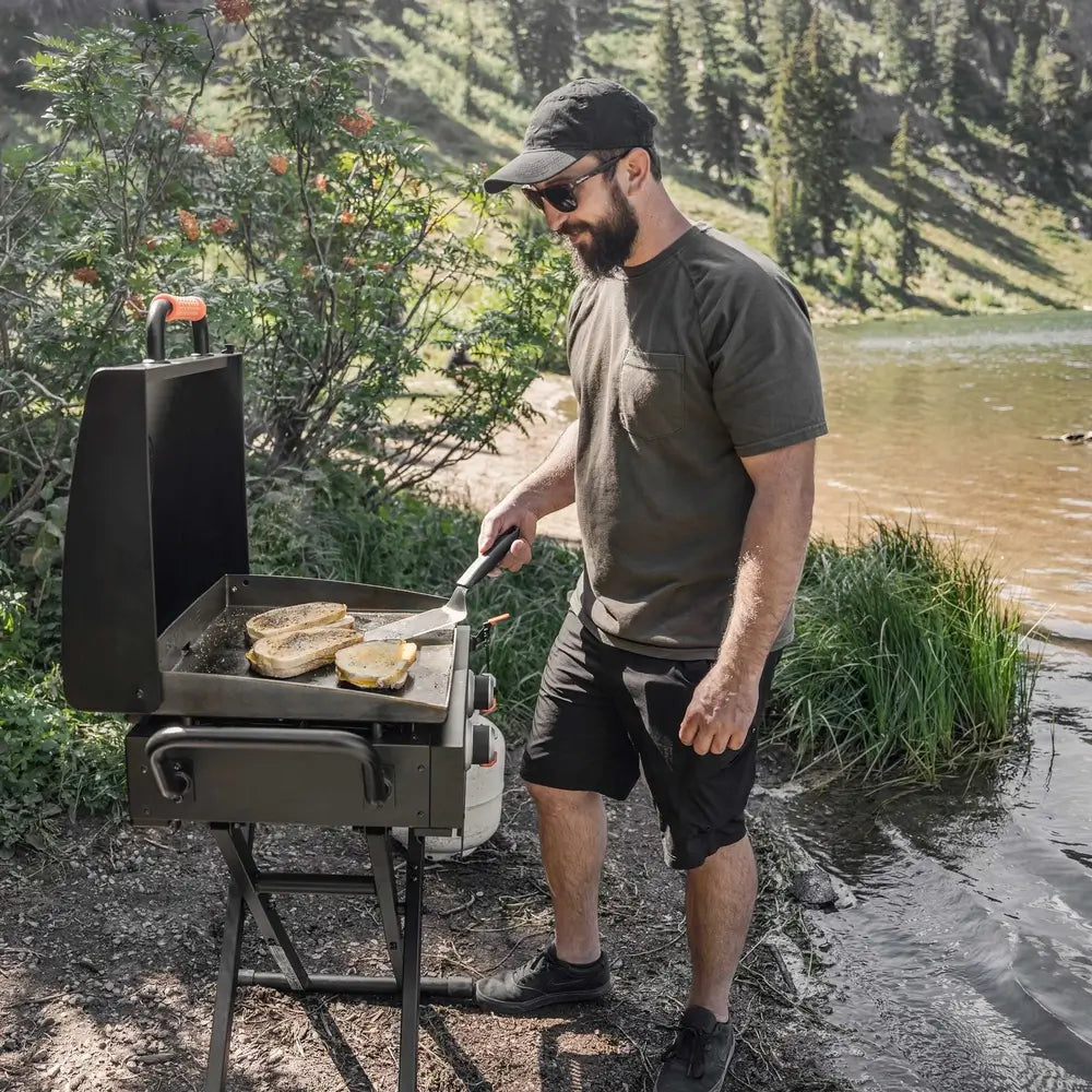 Man using Blackstone 22” outdoor portable flat top griddle to cook sandwiches near scenic lake