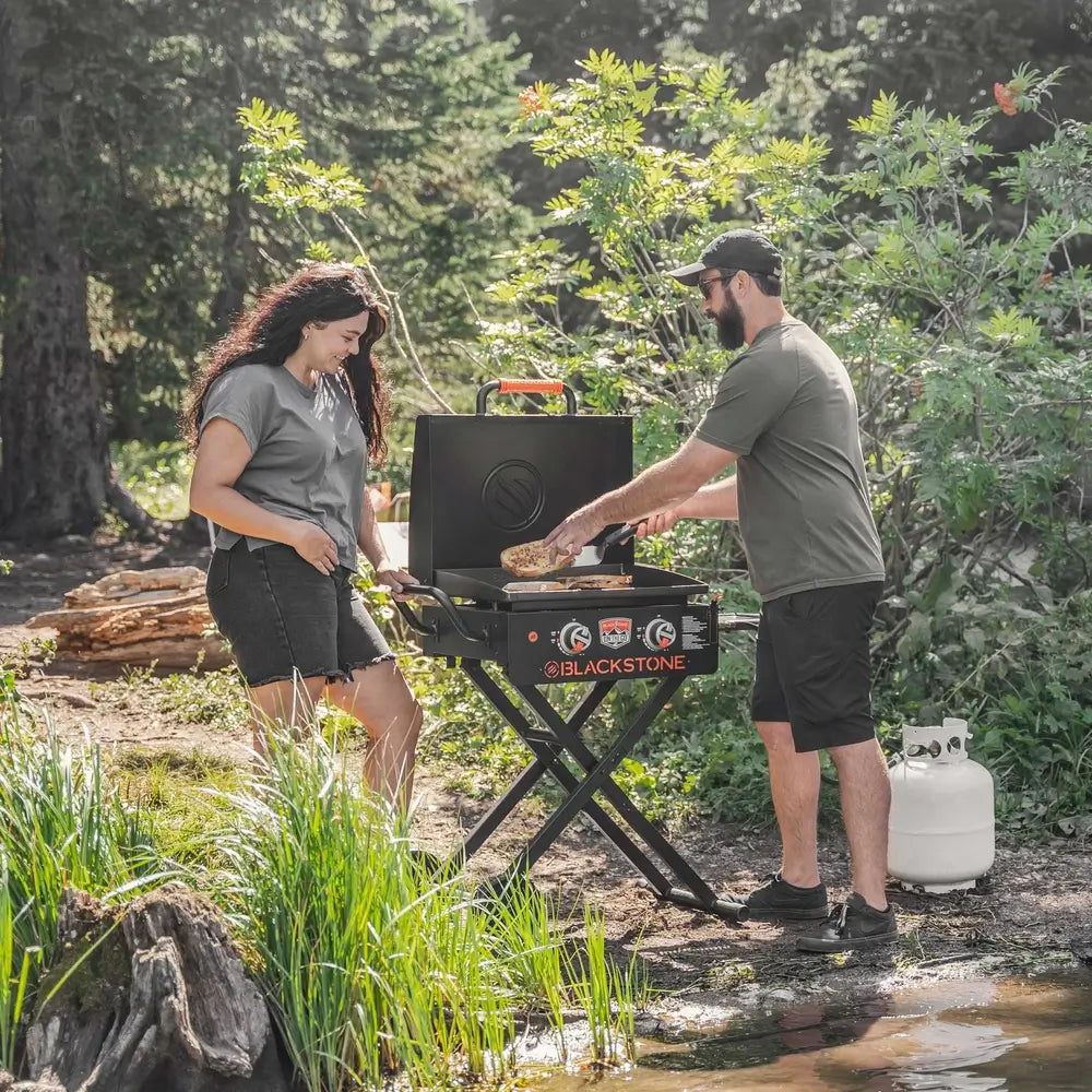 Blackstone 22-inch gas griddle with hood grilling food at forest campsite with propane tank setup