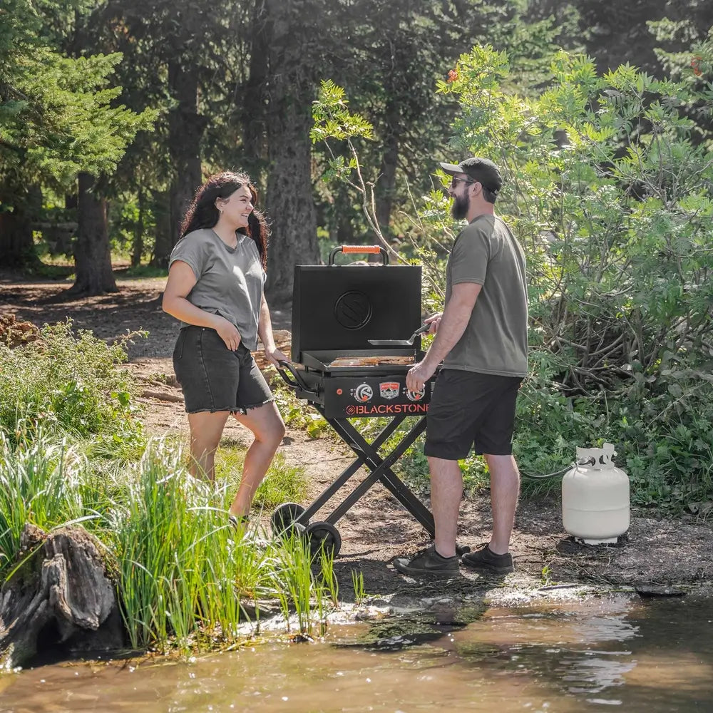 Portable Blackstone propane griddle on scissor cart used for outdoor camping BBQ by a forest lake
