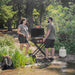 Portable Blackstone propane griddle on scissor cart used for outdoor camping BBQ by a forest lake