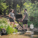 Portable Blackstone propane griddle on scissor cart used for outdoor camping BBQ by a forest lake