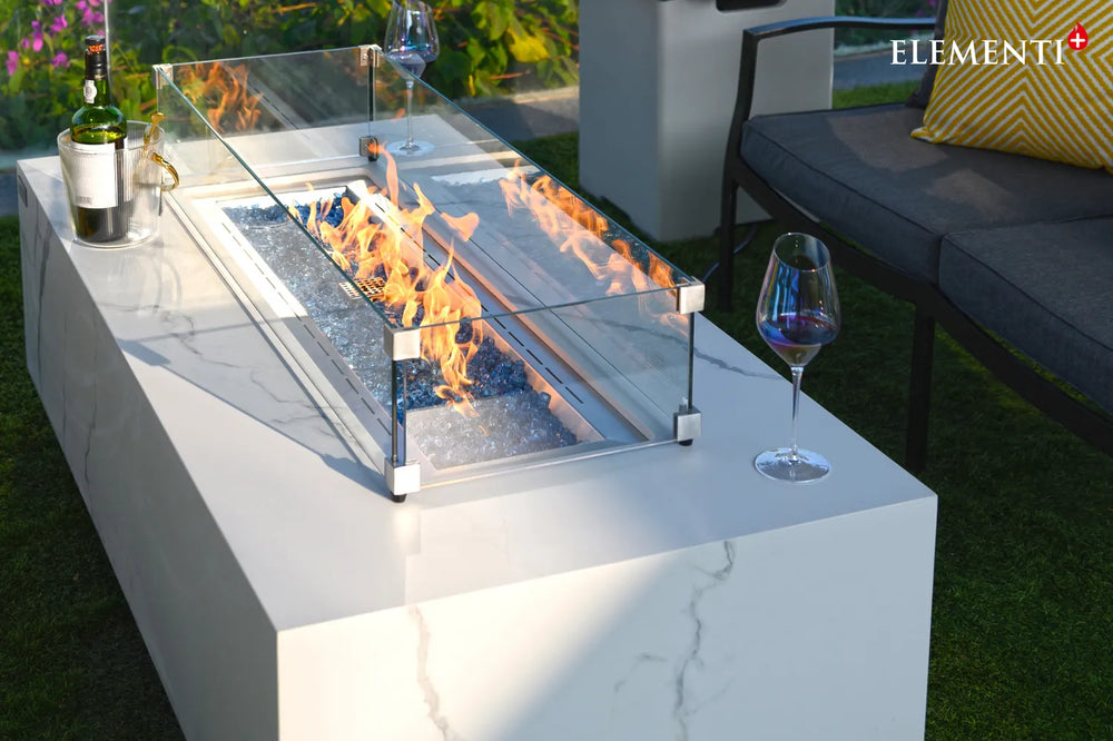 Close-up of the Carrara Bianco fire table's square tempered glass wind guard protecting a vibrant flame over blue fire glass rocks.