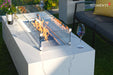 Close-up of the Carrara Bianco fire table's square tempered glass wind guard protecting a vibrant flame over blue fire glass rocks.