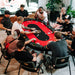 Group of players gathered around the Triton 90-inch premium 10 player folding poker table with red felt, black center lane, padded armrests, and built-in cup holders.