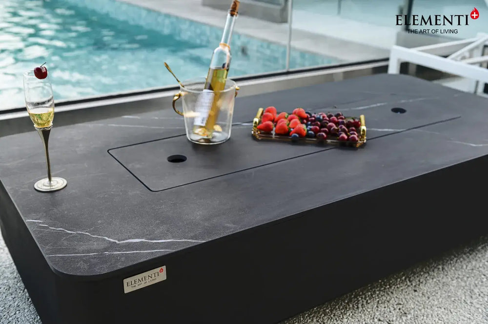 A close-up detail of the fire pit table used as a coffee table. The burner is covered by a matching lid, and a bottle of wine in a bucket, a glass of champagne, and a tray of berries are placed on the marble-textured surface.