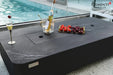 A close-up detail of the fire pit table used as a coffee table. The burner is covered by a matching lid, and a bottle of wine in a bucket, a glass of champagne, and a tray of berries are placed on the marble-textured surface.