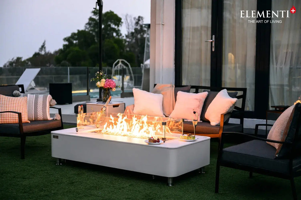 An outdoor evening scene featuring the white marble-effect fire pit table as the centerpiece of a seating area. Champagne glasses and small appetizer plates are placed on the table's edge near the dancing flames.
