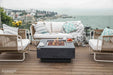 Wide view of a coastal patio with an Elementi gas fire pit, comfortable outdoor seating, and a scenic ocean background.