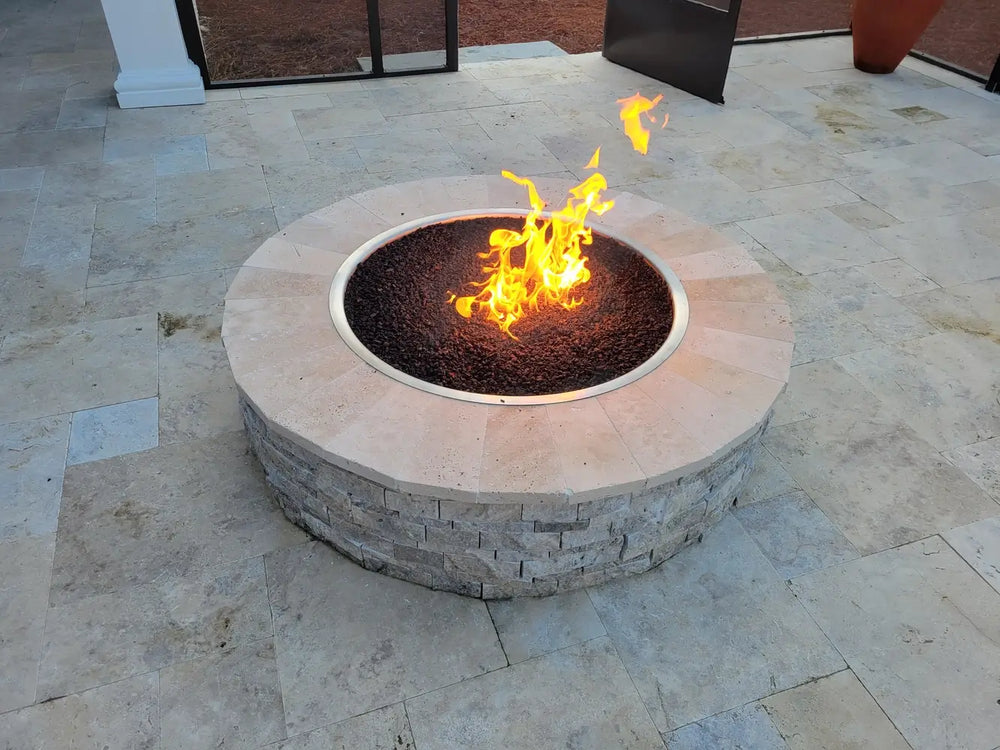 A Travertine TessaRai 46" Round Gas Fire Pit with active flames and black lava rock media installed on a modern light-colored stone patio.
