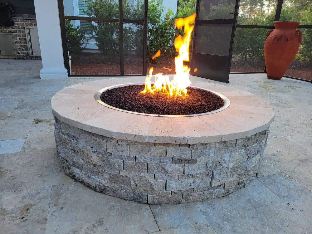Side profile of the Travertine TessaRai 46" Round Gas Fire Pit showing the textured stone base and high-quality burner flames in an outdoor living space.