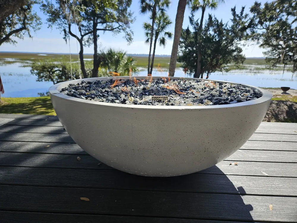 Travertine TessaRai 40 inch Patio Fire Pit Kit with black and clear fire glass on waterfront deck in daylight