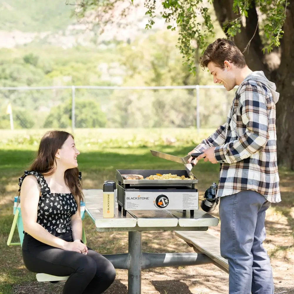 Couple enjoying outdoor cooking with the Blackstone 17" stainless steel tabletop griddle at a park, perfect for portable grilling and campsite meals.