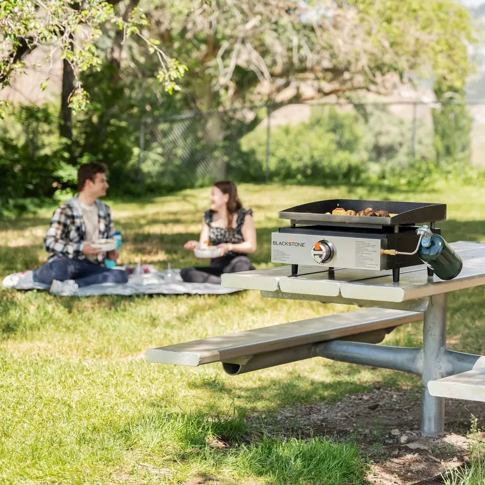 Blackstone 17" tabletop griddle grilling in a scenic outdoor picnic setting with a couple in the background, ideal for portable outdoor cooking and tailgating.