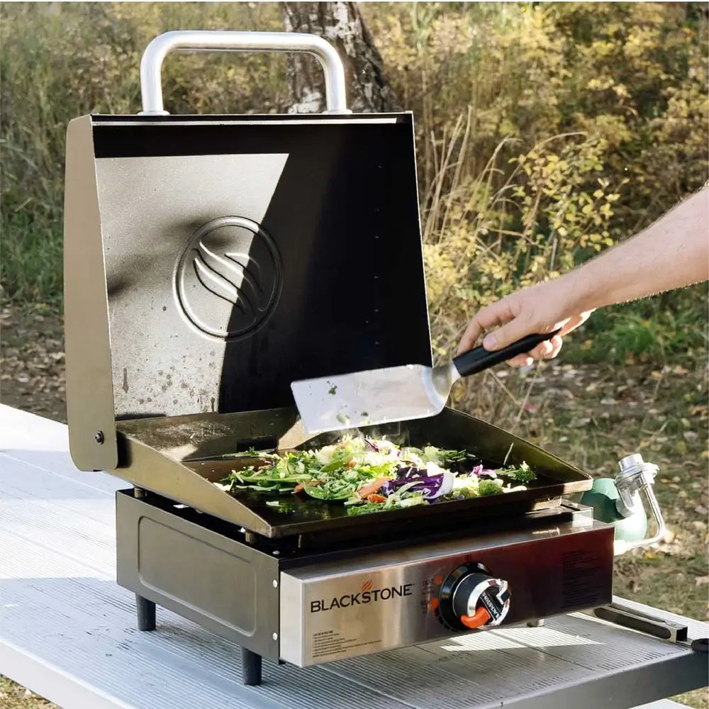 Blackstone 17-inch tabletop gas griddle in action, cooking vegetables with lid open in outdoor campsite setting