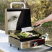 Blackstone 17-inch tabletop gas griddle in action, cooking vegetables with lid open in outdoor campsite setting