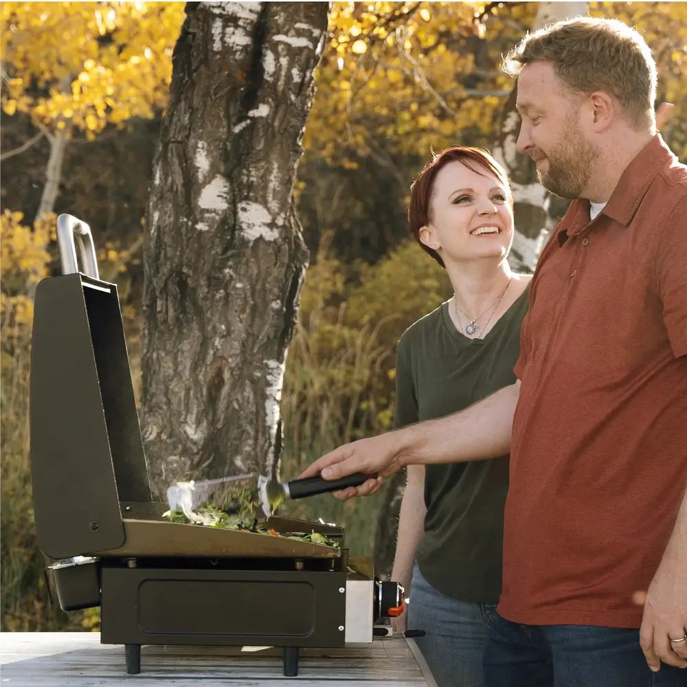 Happy couple cooking on Blackstone 17-inch griddle with hood in a scenic outdoor setting during fall
