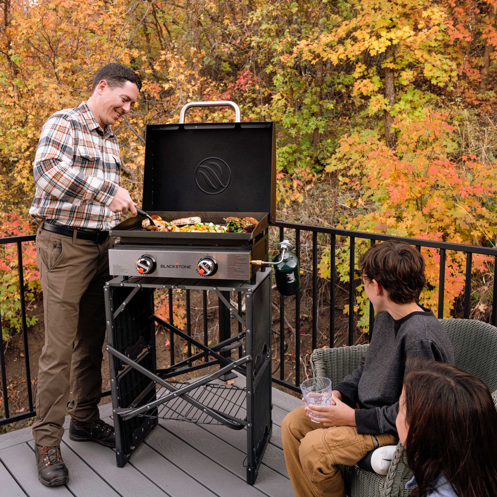 Family grilling on Blackstone 22" portable propane griddle with hood on wooden deck in autumn backdrop