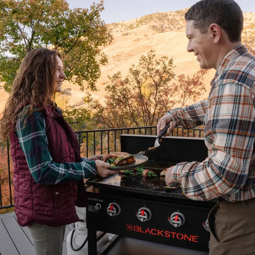 Man serving grilled food from Blackstone stainless steel griddle with hood to woman on patio – high-performance griddle for home chefs