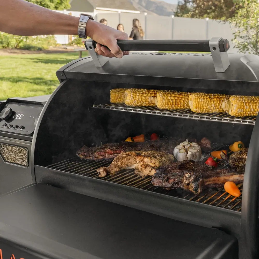Blackstone 900 Pellet Grill open with corn and smoked meats grilling on upper and lower racks in backyard barbecue setting