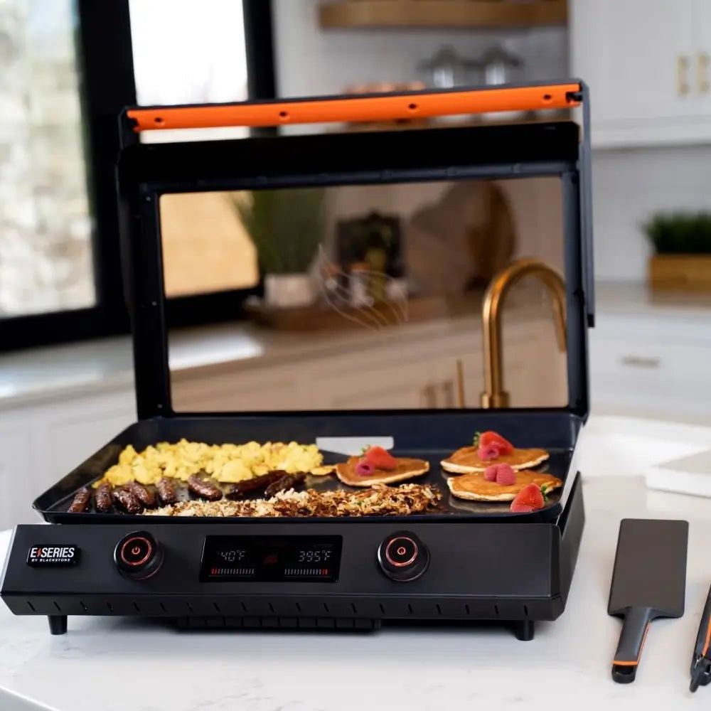 Blackstone 22-Inch E-Series Electric Griddle with glass lid open, cooking breakfast items including sausages, hash browns, and pancakes
