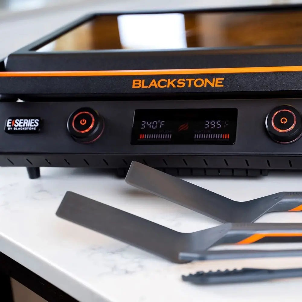 Blackstone E-Series electric griddle with dual spatulas and tongs on counter, showing precision digital controls