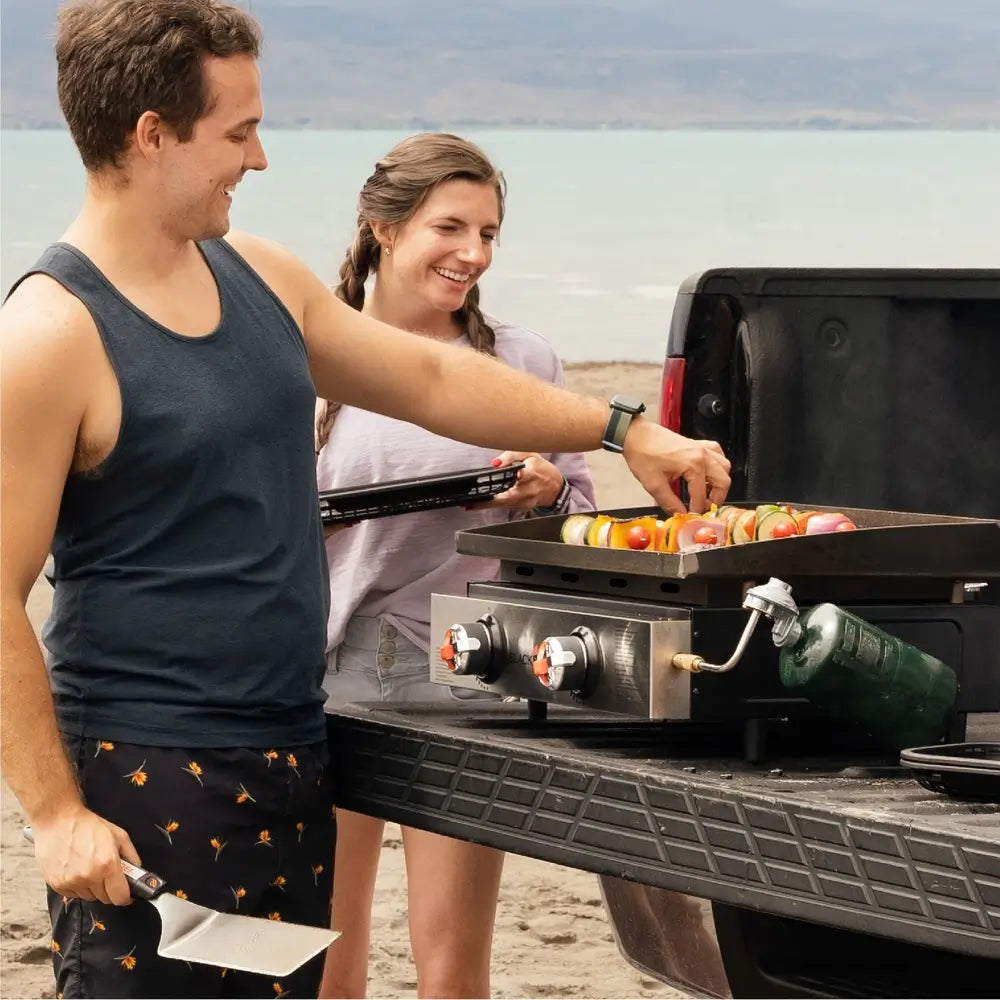 Couple Grilling on Blackstone 22-Inch Propane Tabletop Griddle During Beach Tailgate Picnic