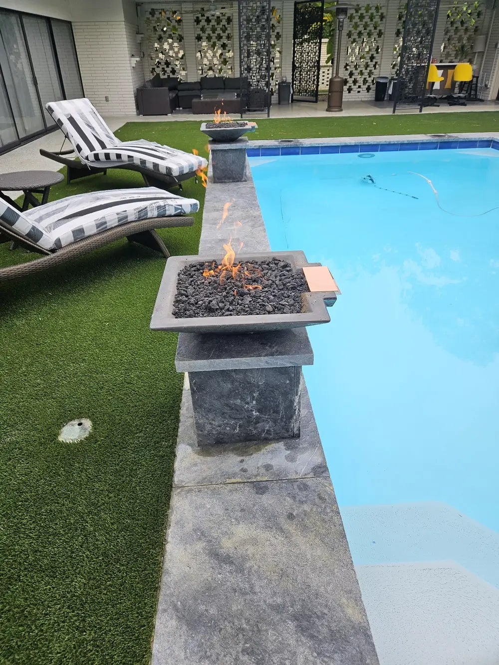 Travertine TessaRai Square Fire/Water Bowl Kit on dark stone pedestal beside pool with turf deck and striped loungers