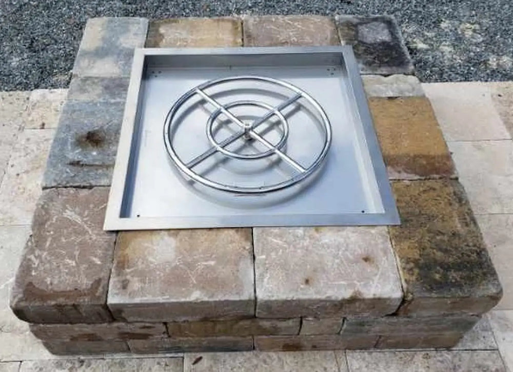 Top-down view of a stainless steel square fire pit pan and double-ring burner kit installed in earth-toned rustic stone pavers.