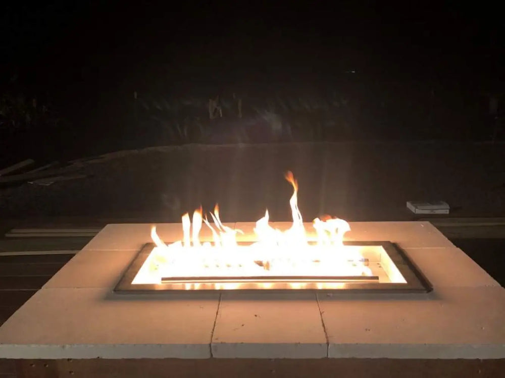 Linear square gas fire pit burner glowing at night with a white stone tile surround and clean modern aesthetic.