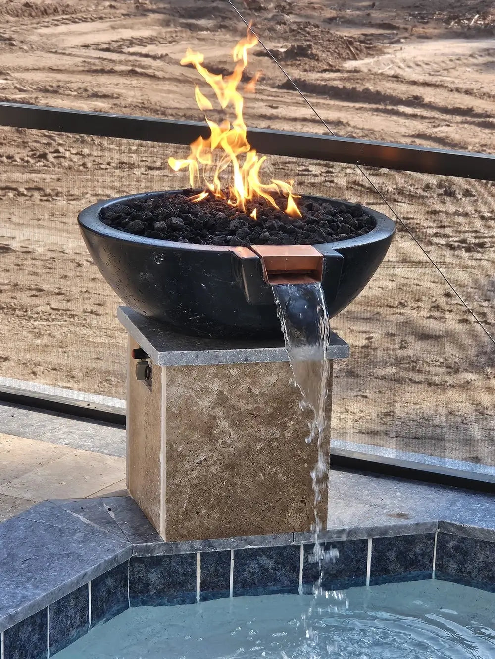 Travertine TessaRai fire and water bowl beside luxury pool with lava rock flames and cascading spillway.