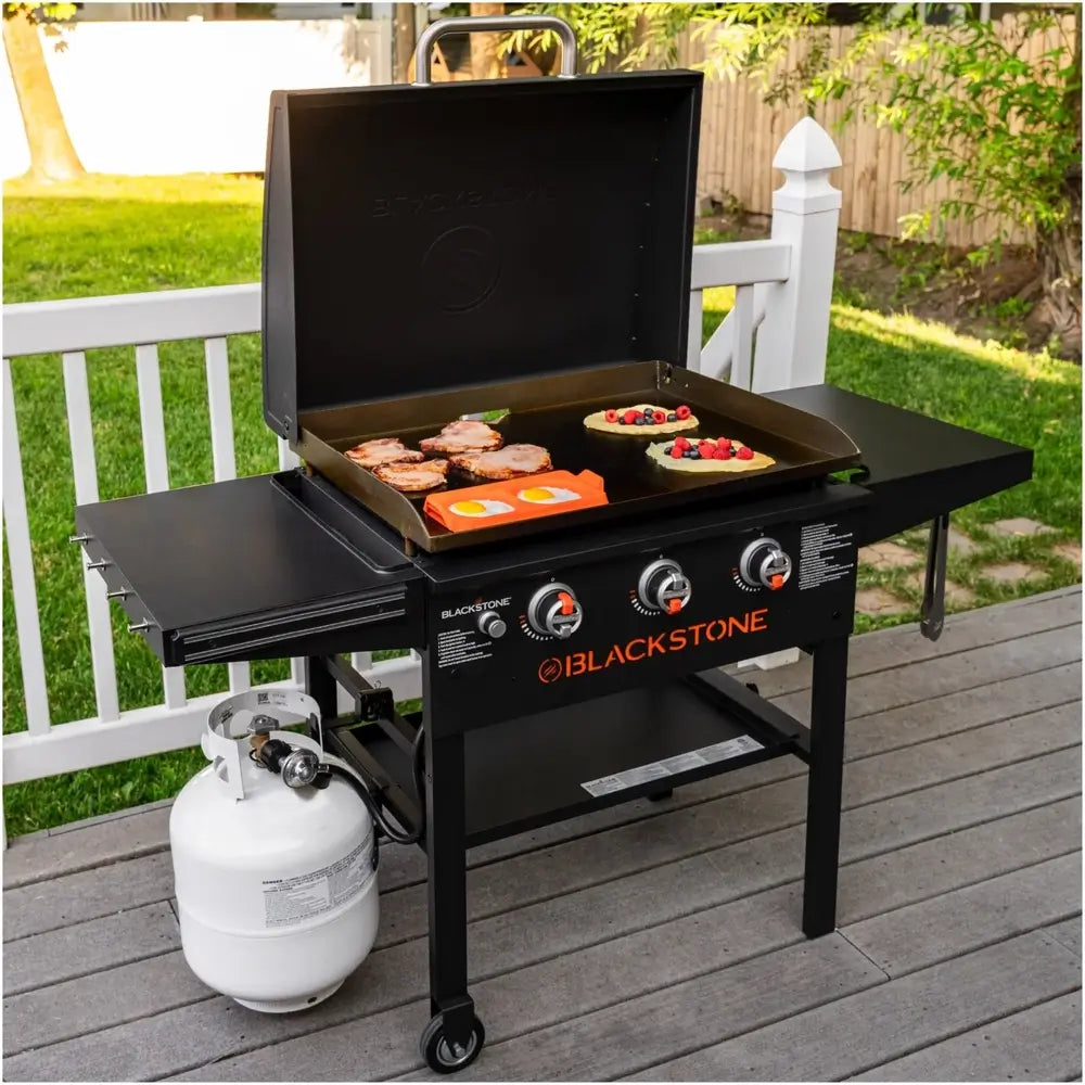 Blackstone propane griddle cooking pancakes, sausage, and eggs with protective hood open on wooden deck
