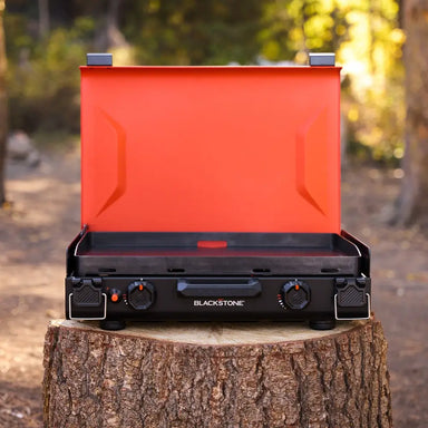 Blackstone 20"x14" portable camping griddle with bright orange hood open, set on a tree stump in the forest, ideal for outdoor cooking.