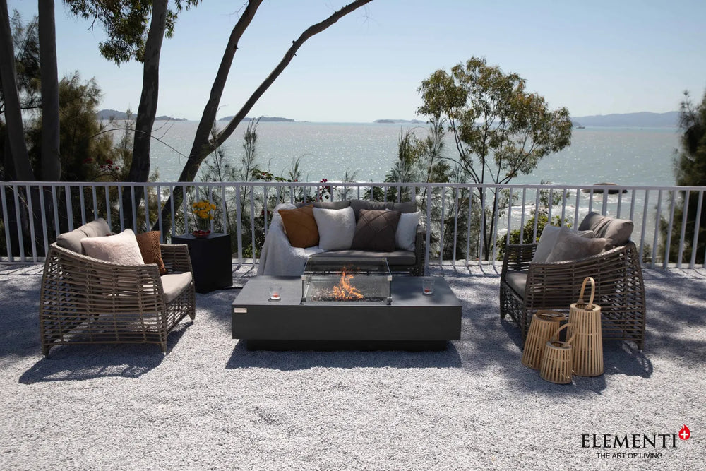 Elementi Plus Cannes 60-inch dark gray fire table on patio with ocean view and wicker furniture.