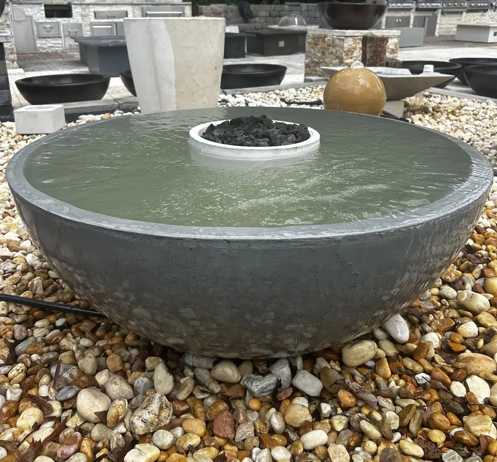 A 40-inch charcoal travertine fire water bowl filled with water on a natural stone pebble base.