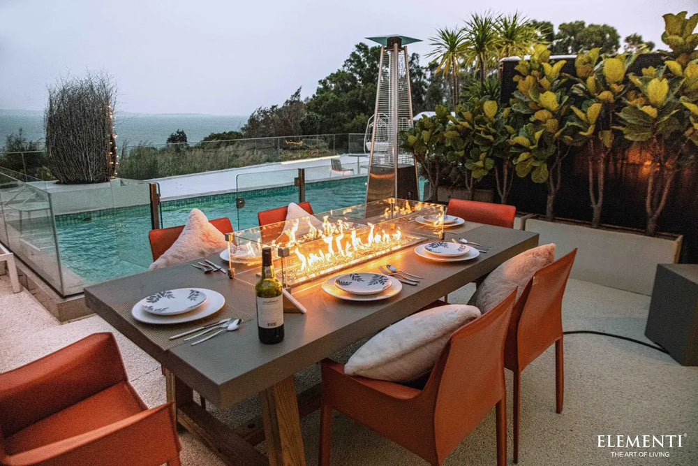 Elementi OFG201 Sonoma Dining Fire Table 45k BTU as a centerpiece on a luxury stone patio with greenery.