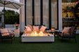 A rectangular white porcelain fire table with grey marble veining sits on an artificial grass patio at dusk. The table features a lit linear fire protected by a glass wind guard, surrounded by a black outdoor sofa and lounge chairs with plush cushions.