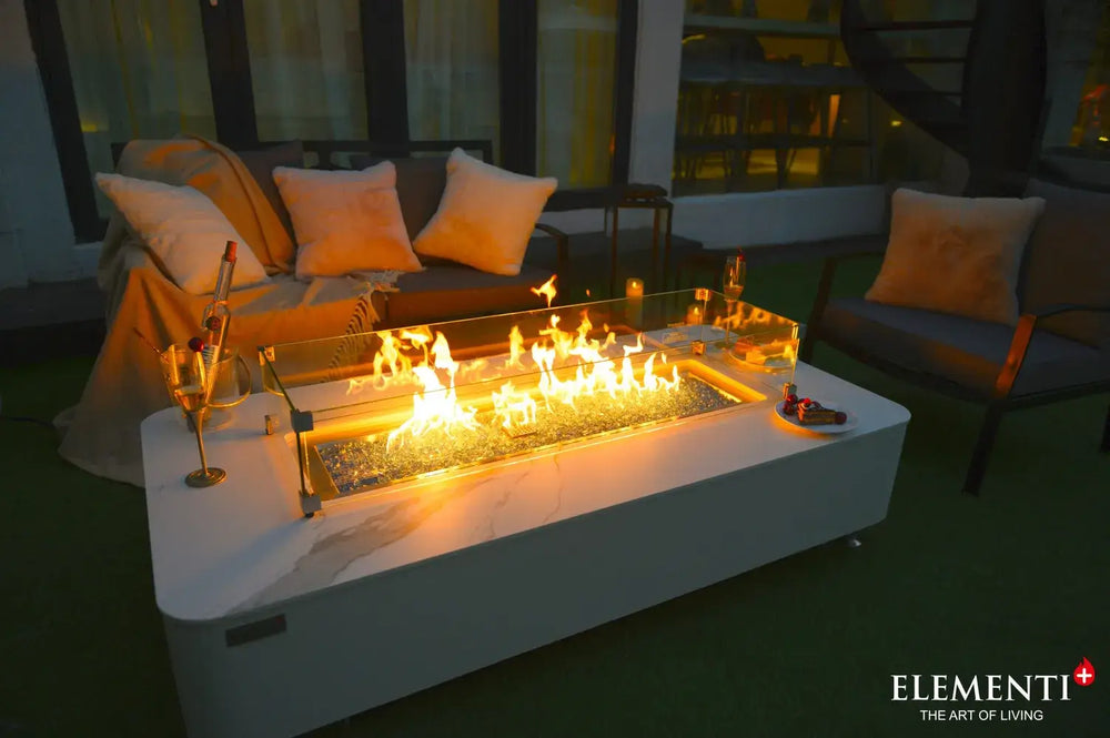 A close-up perspective of the fire table at night, highlighting the orange flames reflected in the clear glass wind guard and the contrasting blue glass fire media.
