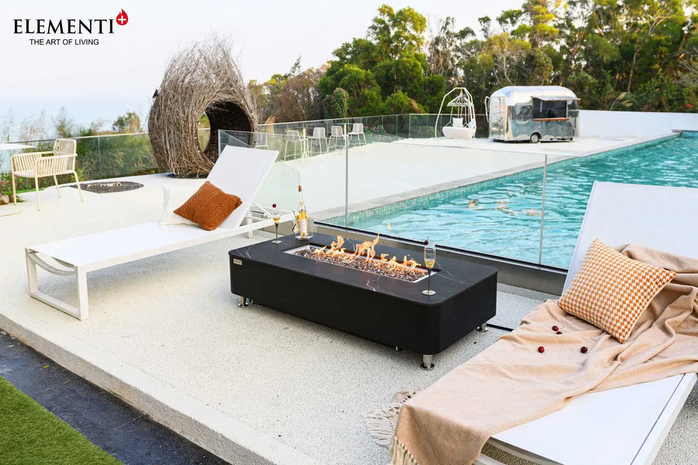 A wide-angle view of a luxury outdoor patio featuring a black fire pit table, white lounge chairs, a unique woven bird’s-nest-style chair, and a vintage silver trailer in the background next to a pool.