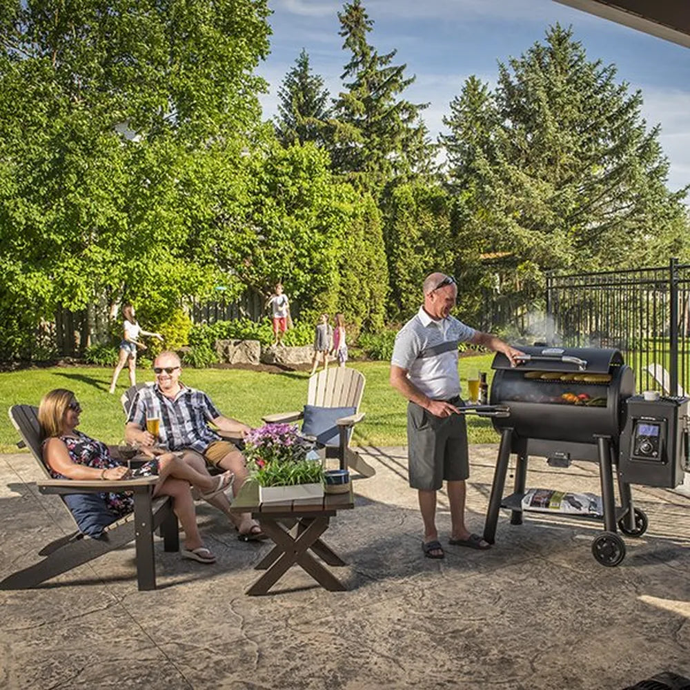 Family relaxing on a backyard patio while a man cooks on a smoking Broil King Regal Pellet 500 WiFi pellet grill, with the pellet smoker featured in an outdoor entertaining setting.