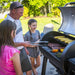 Man grilling with two children beside an open Broil King Regal Pellet 500 WiFi pellet grill in a sunny backyard, with corn, ribs, peppers, and asparagus cooking on the smoker grates.