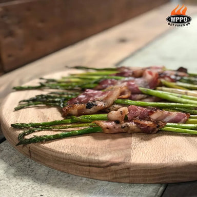 Wood-fired bacon-wrapped asparagus prepared in a WPPO pizza oven on a wooden serving board.